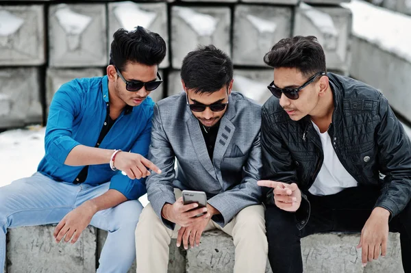 三个随意的年轻印度男人在太阳镜摆在石头块和看着手机 — 图库照片