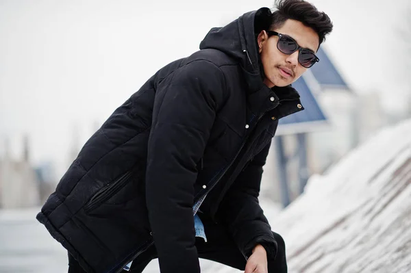 Casual young indian man in black jacket and sunglasses posed at winter day.