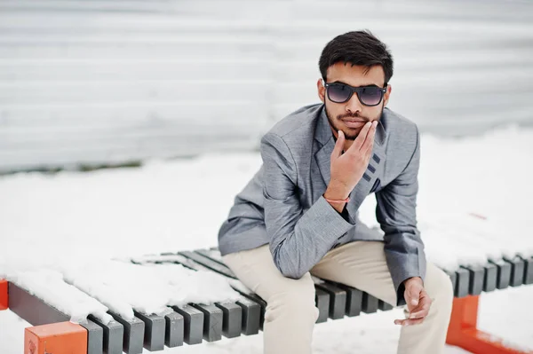 休闲年轻的印度男子在银色夹克和太阳镜摆姿势在冬天的日子 坐在长凳上 — 图库照片