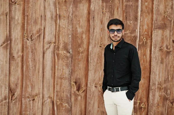 Casual young indian man in black shirt and sunglasses posed against wooden background.