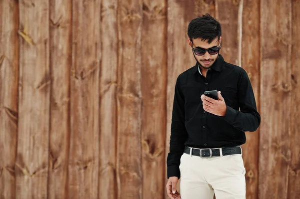 休闲年轻的印度男子在黑色衬衫和太阳镜摆姿势对木背景看着手机 — 图库照片