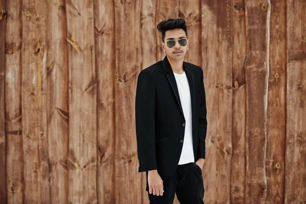 Casual young indian man in black blazer and sunglasses posed against wooden background.