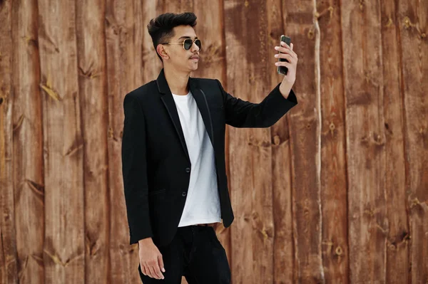 Casual young indian man in black blazer and sunglasses posed against wooden background making selfie at mobile phone.