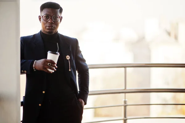 Fashionable african american man in suit and glasses with cup of coffee at hands posed on balkony at office.