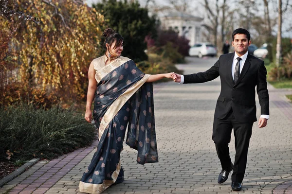 Elegant and fashionable indian friends couple of woman in saree and man in suit walking outdoor and holding hands.