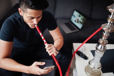 Yakışıklı ve şık Hintli adam siyah odasında oturuyor, nargile sigara ve cep telefonundan arıyor.