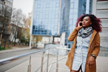 Çekici kıvırcık Afrikalı-Amerikalı kadın kahverengi ceket üstünde hareket eden telefon konuşma modern çok öykülü bina karşı parmaklıklar yakınındaki poz verdi.