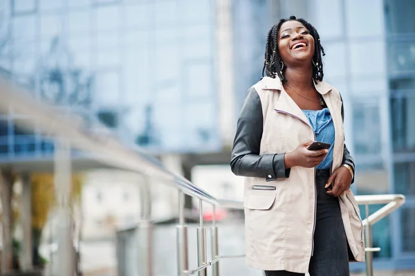 Attractive african american woman with dreads in jacket posed near railings against modern multistory building with mobile phone.