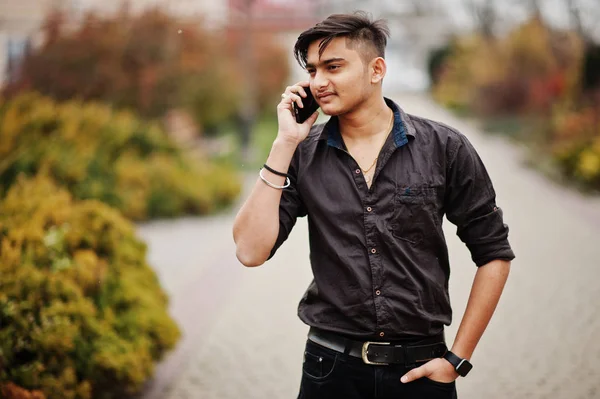Indian man in brown shirt posed outdoor and speaking on mobile phone.