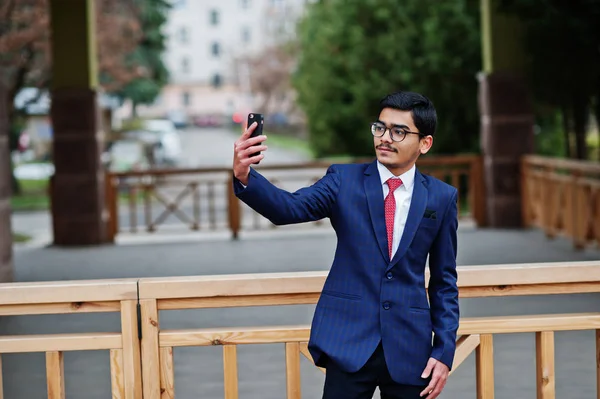 Indian young man at glasses, wear on suit with red tie posed outdoor and making selfie on mobile phone.