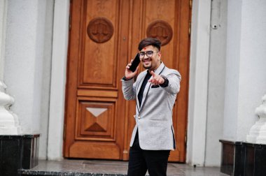 Şık Hintli adam bindi alın ve gözlük, Tarih ile giyim gri takım elbise poz açık kapı binasının, cep telefonu ve gösteri parmak kamera konuşan karşı.