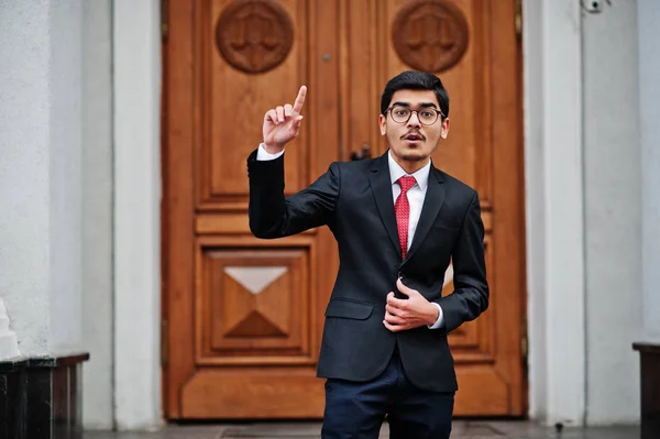 Indian young man at glasses, wear on black suit with red tie posed outdoor against door of building show finger up, he has idea.