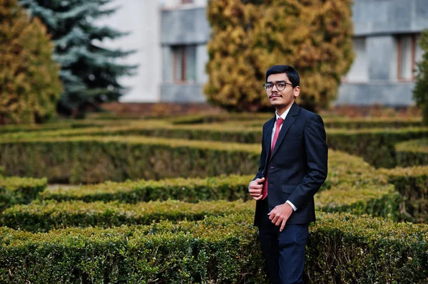 Indian young man at glasses, wear on black suit with red tie posed outdoor.