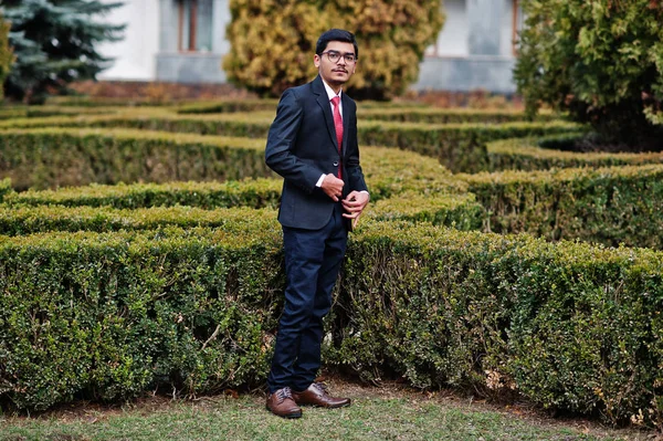 Indian young man at glasses, wear on black suit with red tie posed outdoor.