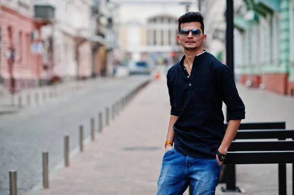 Stylish indian model man in casual clothes, black shirt and sunglasses posed outdoor at street of India.
