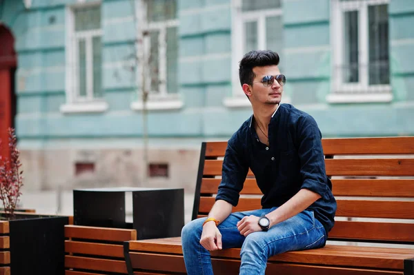 Şık Hint modeli adam rahat kıyafetler, siyah gömlek ve güneş gözlüğü Hindistan ve bankta oturan Street açık poz verdi.