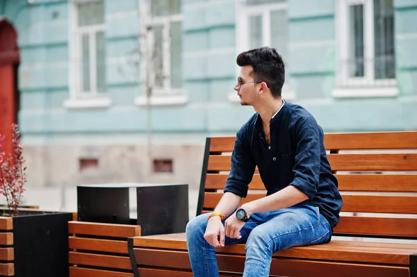Şık Hint modeli adam rahat kıyafetler, siyah gömlek ve güneş gözlüğü Hindistan ve bankta oturan Street açık poz verdi.