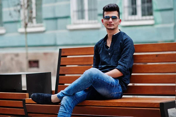 Şık Hint modeli adam rahat kıyafetler, siyah gömlek ve güneş gözlüğü Hindistan ve bankta oturan Street açık poz verdi.