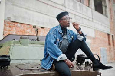 Puro sigara ve btr askeri zırhlı araç karşı yöneltilen Afro-Amerikan erkek kot ceket, bere ve gözlük,.