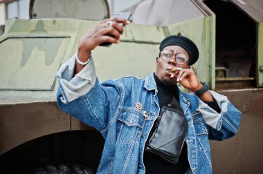 Puro sigara ve btr karşı askeri zırhlı araç, selfie yapım telefon üzerinde yarattığı Afrikalı-Amerikalı adam kot ceket, bere ve gözlük,.