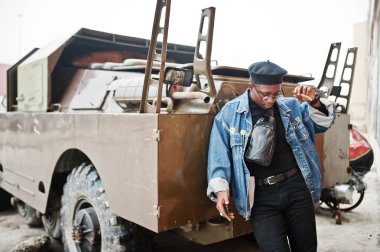 Puro sigara ve btr askeri zırhlı araç karşı yöneltilen Afro-Amerikan erkek kot ceket, bere ve gözlük,.
