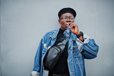 Afrika kökenli Amerikalı adam gri çelik arka plan üzerinde puro sigara kot ceket, bere ve gözlük,.