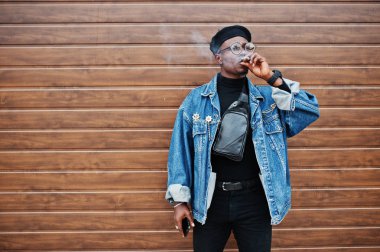 Afrika kökenli Amerikalı adam kahverengi çekim zemin üzerine puro sigara kot ceket, bere ve gözlük,.