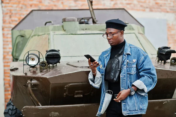 African american man in jeans jacket, beret and eyeglasses, speaking on phone against btr military armored vehicle.