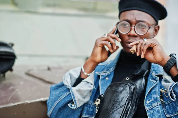 African american man in jeans jacket, beret and eyeglasses, with cigar posed against btr military armored vehicle, and speaking on mobile phone.