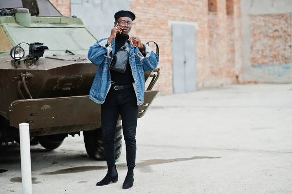 African american man in jeans jacket, beret and eyeglasses, with cigar posed against btr military armored vehicle, and speaking on mobile phone.
