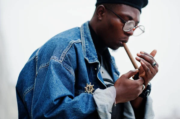 African american man in jeans jacket, beret and eyeglasses, lights a cigar.