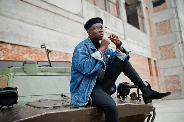 African american man in jeans jacket, beret and eyeglasses, smoking cigar and posed against btr military armored vehicle.