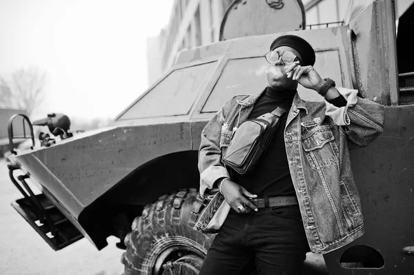 African american man in jeans jacket, beret and eyeglasses, smoking cigar and posed against btr military armored vehicle.
