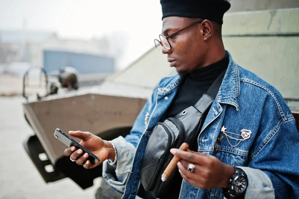 African american man in jeans jacket, beret and eyeglasses, smoking cigar and posed against btr military armored vehicle, with mobile phone at hand.