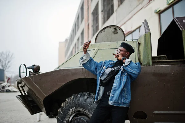 African american man in jeans jacket, beret and eyeglasses, smoking cigar and posed against btr military armored vehicle, making selfie on phone.