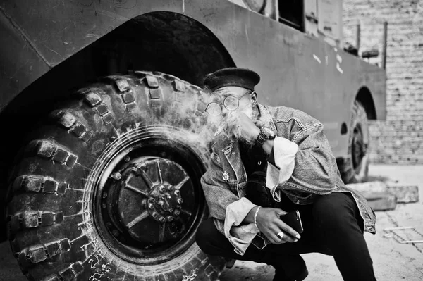 African american man in jeans jacket, beret and eyeglasses, smoking cigar and posed against wheel of btr military armored vehicle.