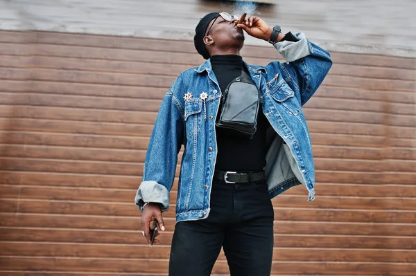 African american man in jeans jacket, beret and eyeglasses, smoking cigar on brown shutter background.