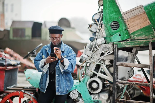 African american man in jeans jacket, beret and eyeglasses, smoking cigar and posed against old retro vehicle and looking at phone.