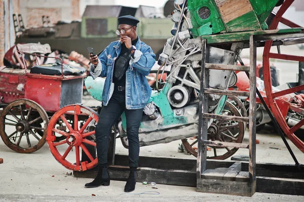 African american man in jeans jacket, beret and eyeglasses, smoking cigar and posed against old retro vehicle and looking at phone.
