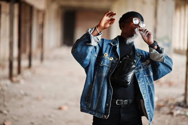 African american man in jeans jacket, beret and eyeglasses, smoking cigar at abandoned factory.