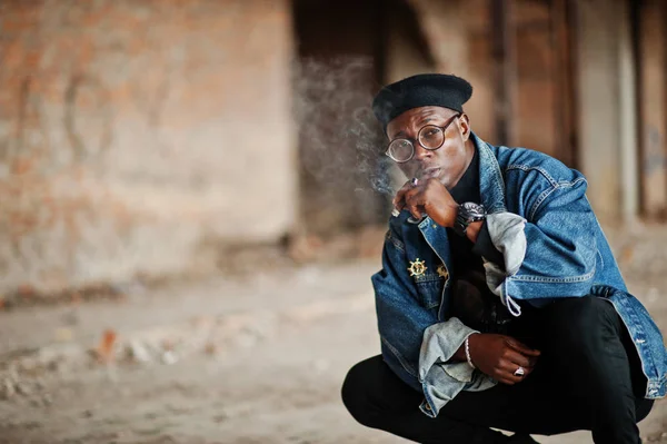 African american man in jeans jacket, beret and eyeglasses, smoking cigar at abandoned factory.