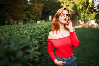 Gözlük, çekici redhaired kadında giymek Kırmızı bluz ve kot etek yeşil park adlı poz.