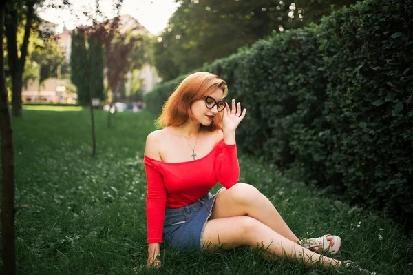 迷人的红头发的妇女在眼镜 穿红色上衣和牛仔裤裙子摆在绿色公园 — 图库照片