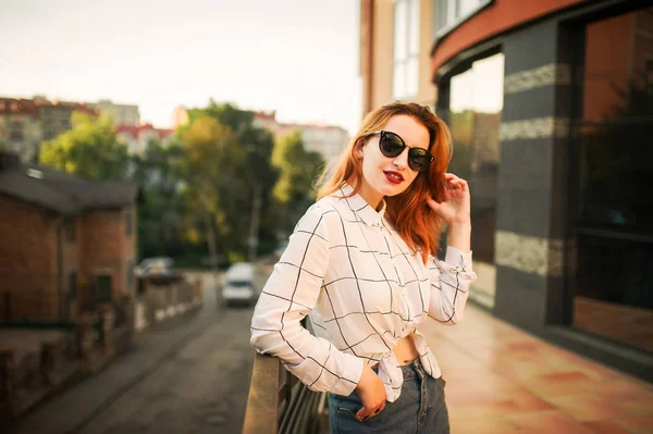 迷人的红头发的女人戴太阳镜 穿在白色上衣摆姿势反对现代建筑 — 图库照片
