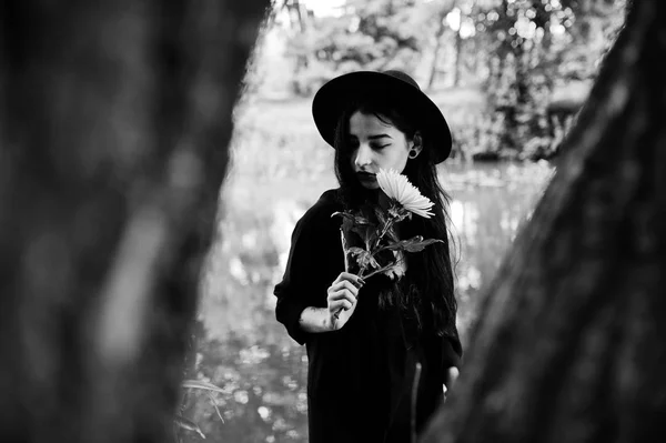 Sensual girl all in black, red lips and hat. Goth dramatic woman hold white chrysanthemum flower at hand against silent lake. Black and white portrait.