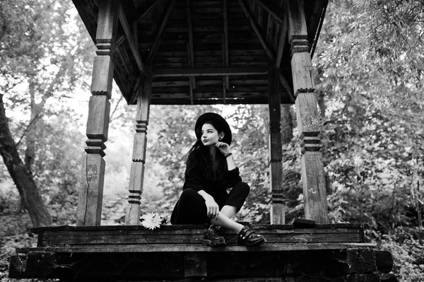 Sensual girl all in black, red lips and hat. Goth dramatic woman sitting on wooden arch at forest. Black and white portrait.