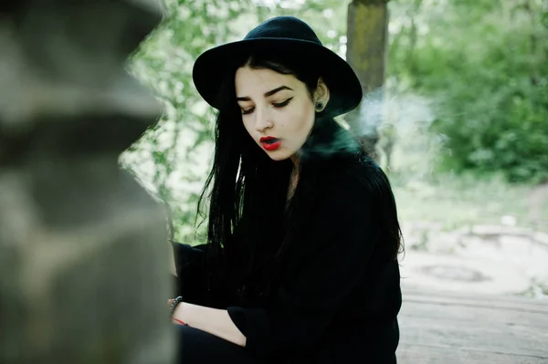 Sensual smoker girl all in black, red lips and hat. Goth dramatic woman smoking thin cigarette.