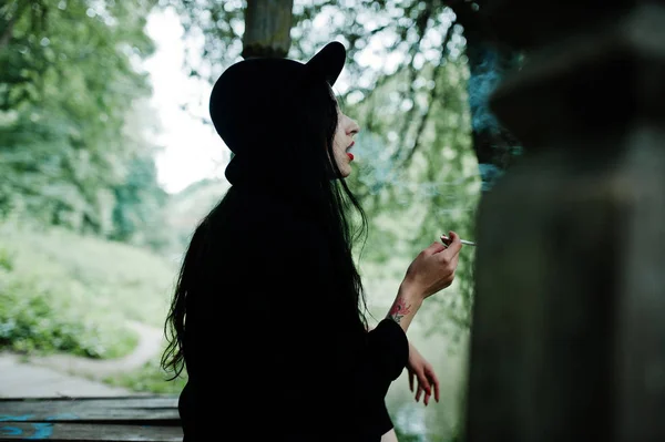 Sensual smoker girl all in black, red lips and hat. Goth dramatic woman smoking thin cigarette.