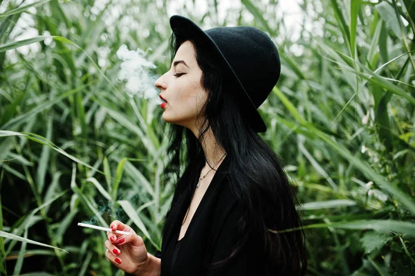 Sensual smoker girl all in black, red lips and hat. Goth dramatic woman smoking on common reed.