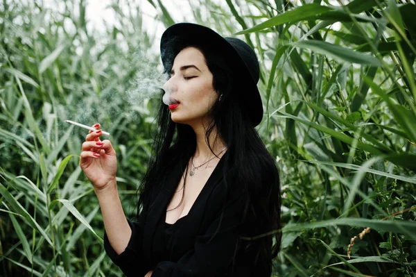 Sensual smoker girl all in black, red lips and hat. Goth dramatic woman smoking on common reed.